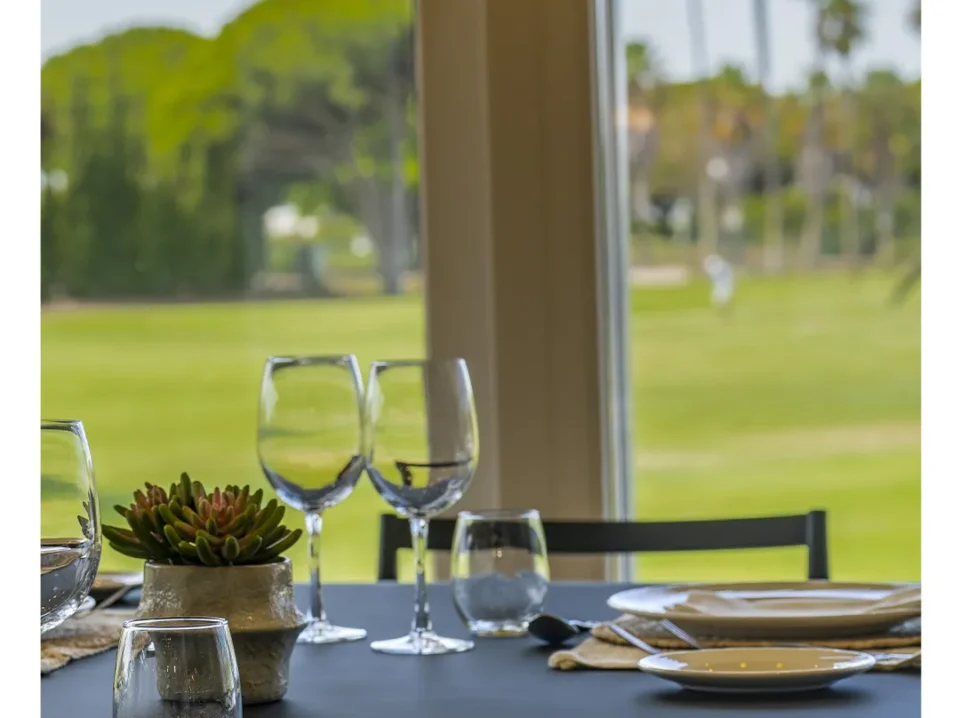Mesa vestida junto al ventanal del restaurante con vistas a los fairways del Real Novo Sancti Petri, con pinar y palmeras al fondo