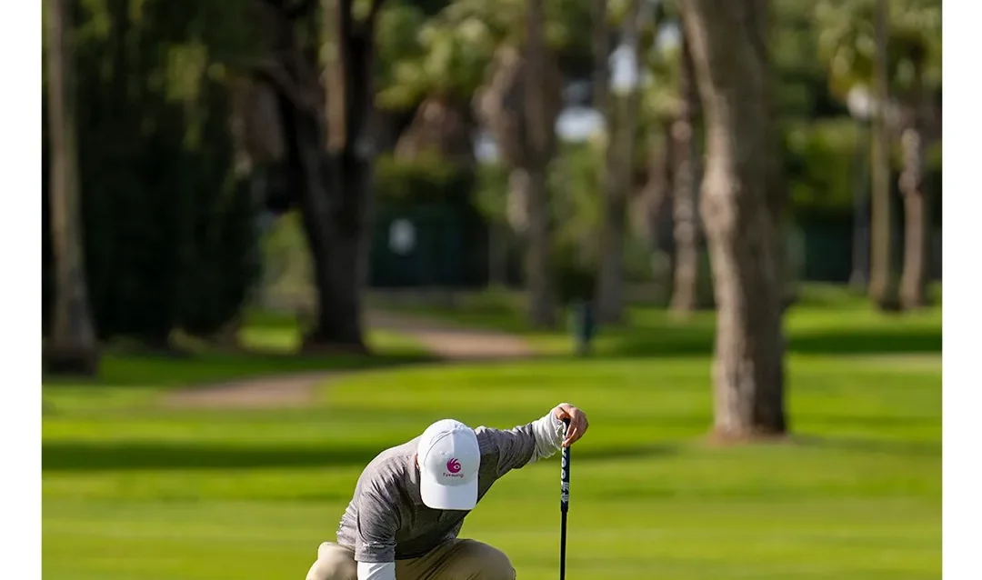 arreglar-piques-en-el-green Golfista agachado arregla piques en el green de Real Novo Sancti Petri con el putter apoyado, entre pinos y palmeras