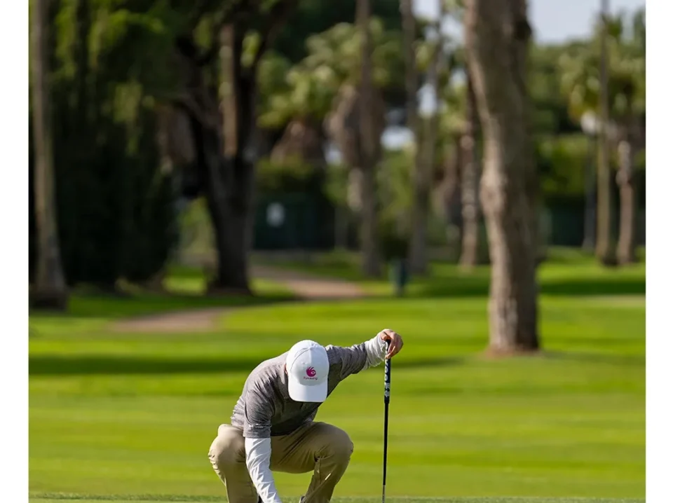 arreglar-piques-en-el-green Golfista agachado arregla piques en el green de Real Novo Sancti Petri con el putter apoyado, entre pinos y palmeras