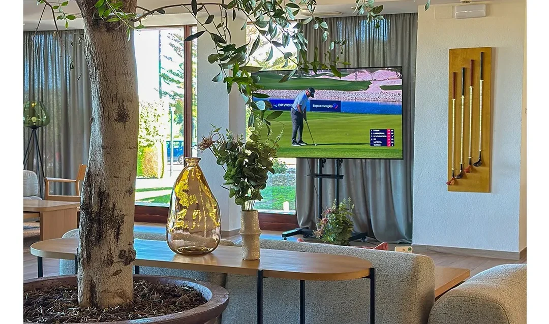 casa-club-de-novo-sancti-petri Salón de la casa club de Novo Sancti Petri con un olivo en maceta, sofás y una pantalla emitiendo un torneo de golf en directo