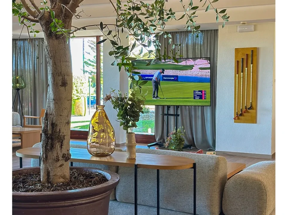 casa-club-de-novo-sancti-petri Salón de la casa club de Novo Sancti Petri con un olivo en maceta, sofás y una pantalla emitiendo un torneo de golf en directo